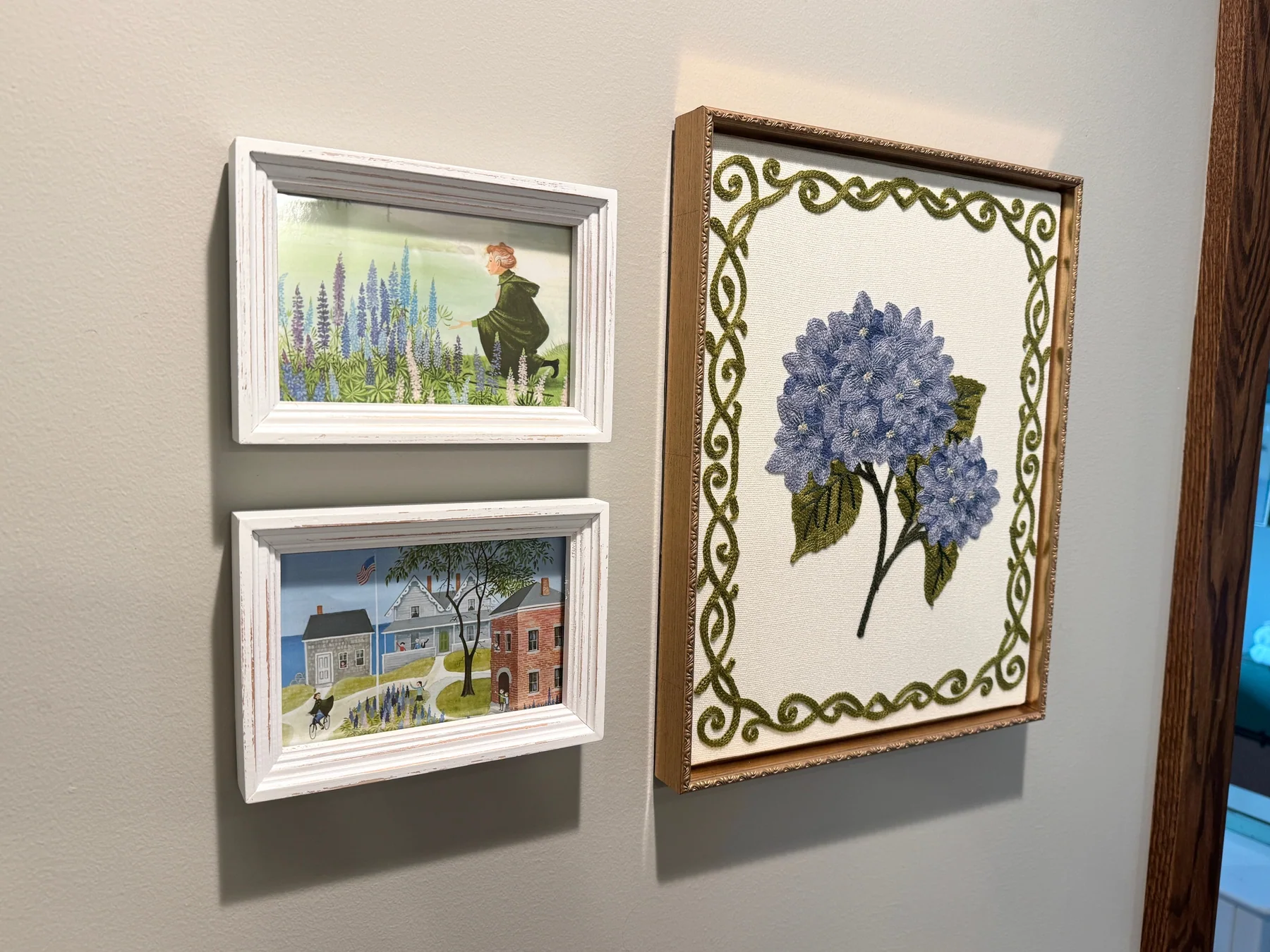 Two framed scenes from Miss Rumphius beside a framed floral needlework piece at Grey Cloud Island